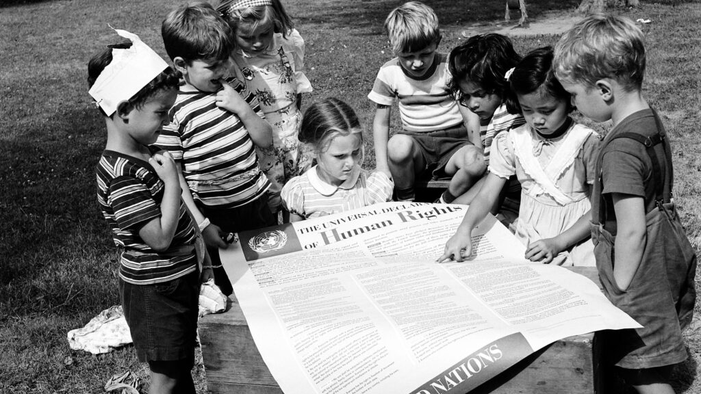 Sort-hvitt-foto av barn på en plen som holder opp og peker på et stort ark med skrift.