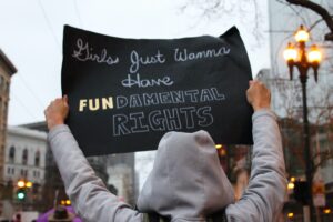 Demonstrant i grå hettejakke som holder opp en svart plakat med skriften "Girls just Wanna Have FUNDAMENTAL RIGHTS"