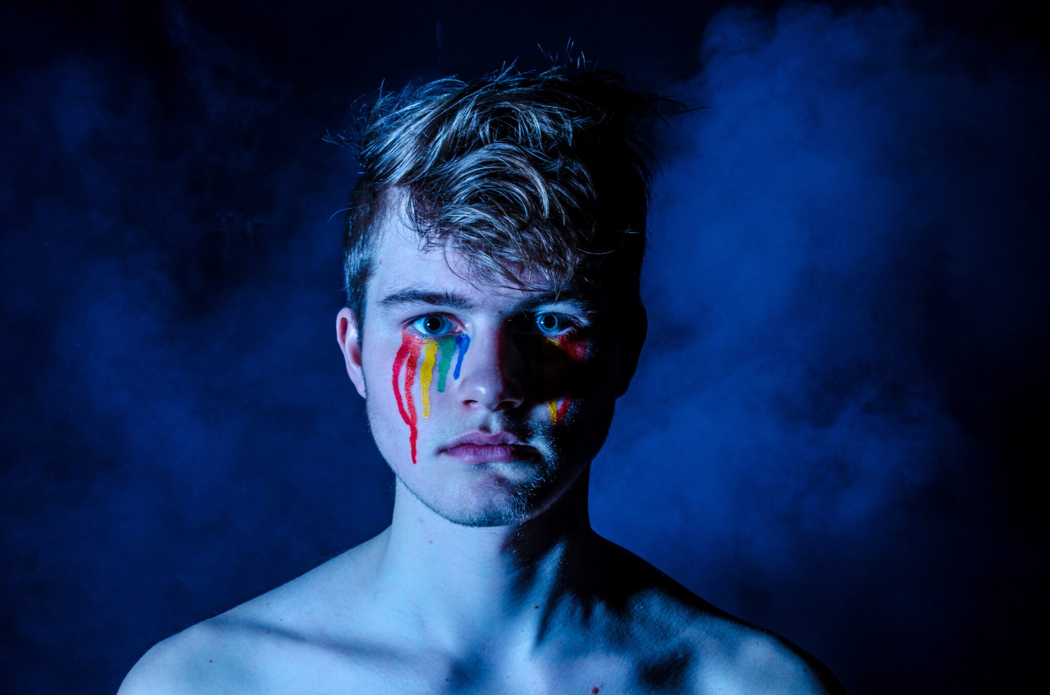 Ansikt på ung mann i dunkel, blå belysning som er malt med tårer i regnbueflaggets farger